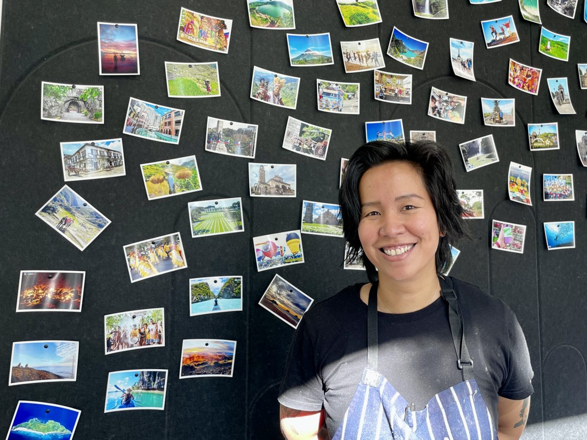 A woman with short hair, wearing an apron stands in front of a pin board covered in colourful postcards.