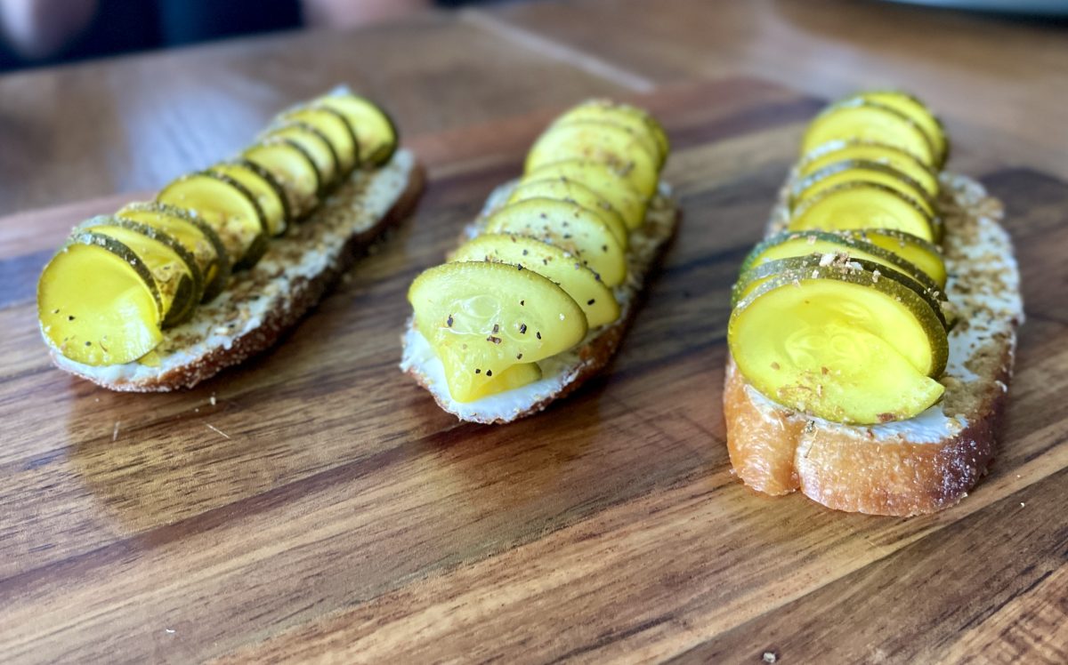 Three pieces of baguette sliced thinly on a wooden board and topped with labneh and zucchini cut in a spiral with a sprinkling of dukkah.