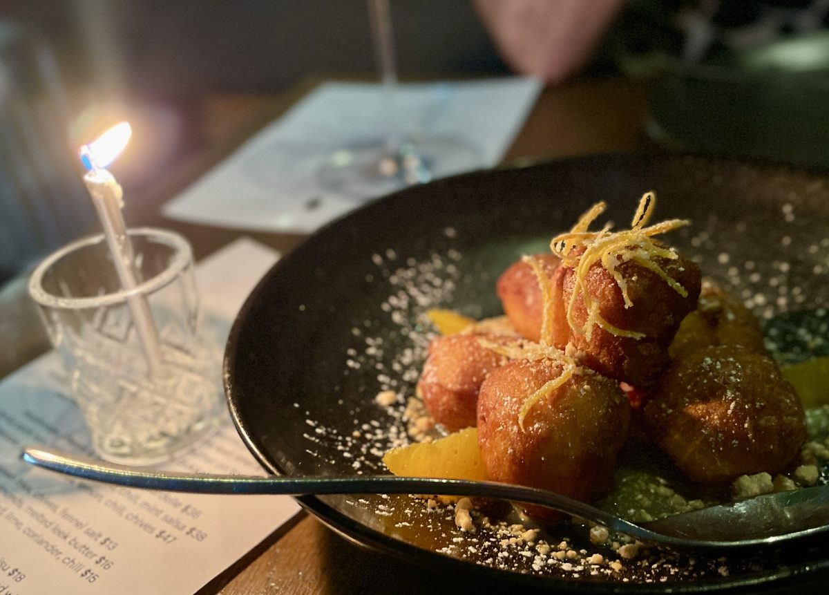 A plated with a stack of small, round doughnuts garnished with orange. A birthday candle in a shot glass sits next to the plate