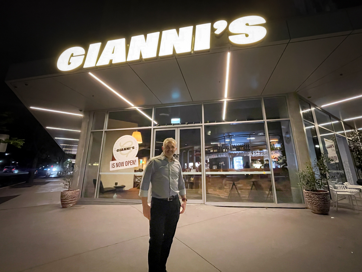 A man stands in front of a restaurant. The name 'Gianni's' is lit up, and the street is dark around it.