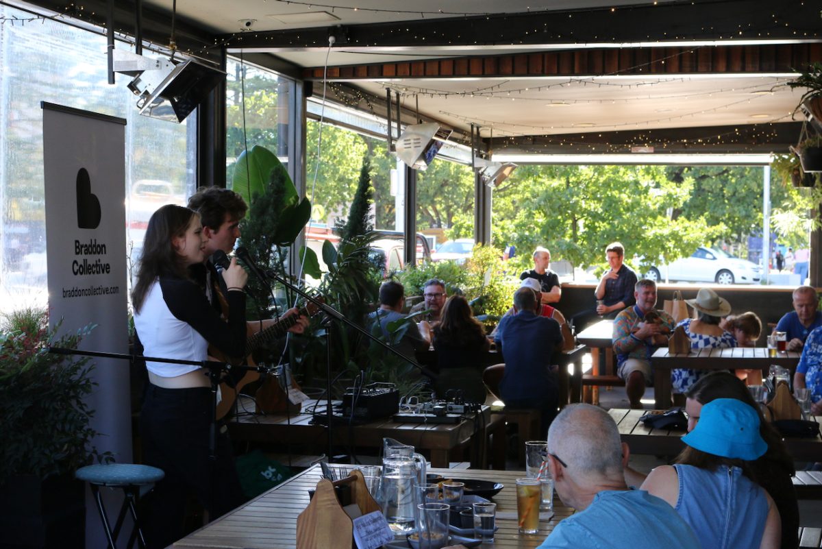 Two young people sing and play guitar at a pub, there is a crowd of people sitting at tables enjoying the music.