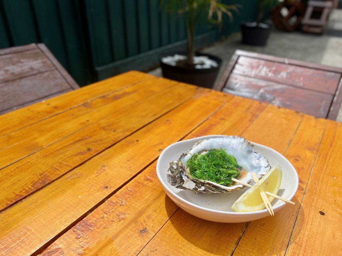 A very large oyster topped with green wakame seaweed sits on a plate next to a wedge of lemon. 